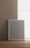 A blank gray picture frame with a white border is leaning against a beige wall on a wooden floor. Soft natural light is coming through a large window on the right, creating a calm and minimalistic atmosphere.