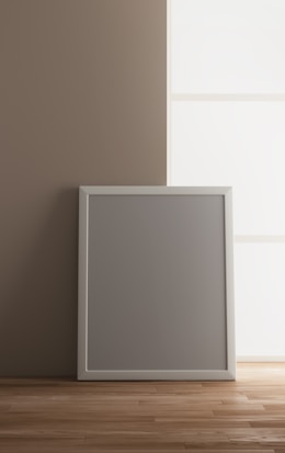 A blank gray picture frame with a white border is leaning against a beige wall on a wooden floor. Soft natural light is coming through a large window on the right, creating a calm and minimalistic atmosphere.