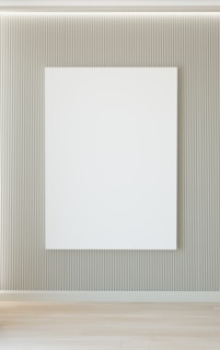 A minimalist interior featuring a large blank white canvas on a wall with vertical grooves. The floor is wooden, and there's soft lighting from above.