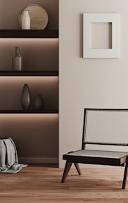 A minimalist interior featuring a wooden chair with a light woven seat and backrest, placed on a wooden floor. On the left, built-in shelves contain decorative objects such as vases, and there is a textured fabric draped at the bottom. A square white frame hangs on the light-colored wall.