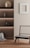 A minimalist interior featuring a wooden chair with a light woven seat and backrest, placed on a wooden floor. On the left, built-in shelves contain decorative objects such as vases, and there is a textured fabric draped at the bottom. A square white frame hangs on the light-colored wall.