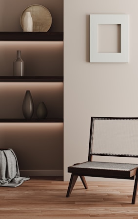 A minimalist interior featuring a wooden chair with a light woven seat and backrest, placed on a wooden floor. On the left, built-in shelves contain decorative objects such as vases, and there is a textured fabric draped at the bottom. A square white frame hangs on the light-colored wall.