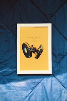 A framed poster with a yellow background depicting abstract black and gold headphones. The title 'Rebellion of Baron' is printed above the headphones. The frame rests on a textured blue fabric surface.