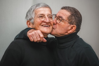 An elderly couple shares a tender and affectionate moment. The man is giving a gentle kiss on the cheek to the woman who is smiling joyfully. Both are wearing glasses and dressed in black turtlenecks. Their expressions convey a deep sense of love and companionship.