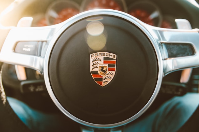 Close-up of a Porsche steering wheel, prominently featuring the Porsche logo in the center. The surrounding area includes chrome accents and visible instrumentation in the background.