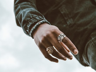 A close-up of a hand wearing several ornamental rings, including one with a skull design and another that appears to be of a lion's face. The person is wearing a green long-sleeved shirt and a beaded bracelet.