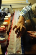 An elderly person's hand arranging pills into a weekly pill organizer. Several prescription medicine bottles are seen on a dark wooden surface. The person is wearing a light blue, patterned sleeve. The background includes a blurred section with various household items.