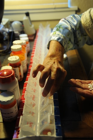 An elderly person's hand arranging pills into a weekly pill organizer. Several prescription medicine bottles are seen on a dark wooden surface. The person is wearing a light blue, patterned sleeve. The background includes a blurred section with various household items.
