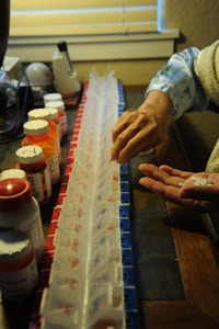 An elderly hand transfers pills from a weekly pill organizer into another hand. The organizer is color-coded and labeled with days of the week. Prescription medication bottles are lined up beside the organizer. The lighting is warm and soft, creating a cozy atmosphere.