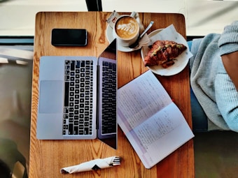 A wooden table with a laptop, notebook with handwritten notes, coffee cup with latte art, and a plate with a croissant. A fork wrapped in a napkin lies beside the laptop, and there appears to be a person wearing a sweater sitting at the table.