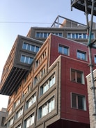 A modern building with an unconventional design, featuring overlapping geometric shapes and varying facade textures. The structure includes sections with gray bricks and red panels, creating a striking contrast. Large windows with white frames are evenly distributed, reflecting the clear sky.