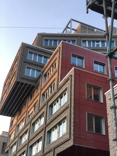 A modern building with an unconventional design, featuring overlapping geometric shapes and varying facade textures. The structure includes sections with gray bricks and red panels, creating a striking contrast. Large windows with white frames are evenly distributed, reflecting the clear sky.