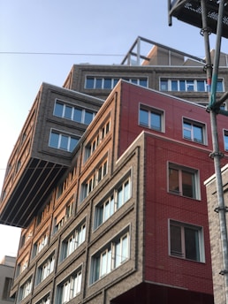 A modern building with an unconventional design, featuring overlapping geometric shapes and varying facade textures. The structure includes sections with gray bricks and red panels, creating a striking contrast. Large windows with white frames are evenly distributed, reflecting the clear sky.