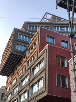A modern building with an unconventional design, featuring overlapping geometric shapes and varying facade textures. The structure includes sections with gray bricks and red panels, creating a striking contrast. Large windows with white frames are evenly distributed, reflecting the clear sky.
