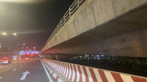 A nighttime scene features a multi-lane roadway beneath a large concrete overpass. The road surface is illuminated by streetlights, and traffic can be seen moving smoothly. A striped barrier runs alongside the road, with direction arrows painted on the pavement. In the distance, illuminated road signs with blue backgrounds and white text guide drivers.