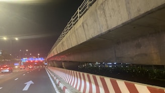 A nighttime scene features a multi-lane roadway beneath a large concrete overpass. The road surface is illuminated by streetlights, and traffic can be seen moving smoothly. A striped barrier runs alongside the road, with direction arrows painted on the pavement. In the distance, illuminated road signs with blue backgrounds and white text guide drivers.