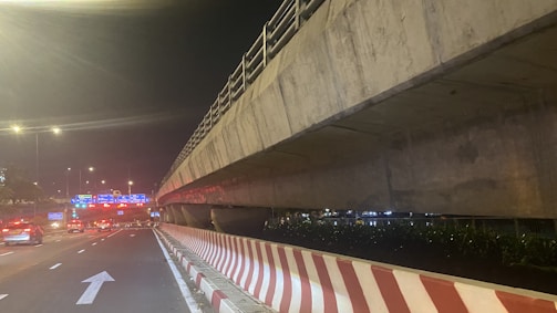 A nighttime scene features a multi-lane roadway beneath a large concrete overpass. The road surface is illuminated by streetlights, and traffic can be seen moving smoothly. A striped barrier runs alongside the road, with direction arrows painted on the pavement. In the distance, illuminated road signs with blue backgrounds and white text guide drivers.
