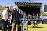 A band is performing on an outdoor stage with several musicians playing instruments, including guitars and drums. Two men in the foreground appear to be managing equipment, possibly part of the event staff. The stage is set up with a black backdrop and surrounded by speakers. The event seems to be taking place in a park or open field, with trees visible in the background.