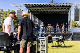 A band is performing on an outdoor stage with several musicians playing instruments, including guitars and drums. Two men in the foreground appear to be managing equipment, possibly part of the event staff. The stage is set up with a black backdrop and surrounded by speakers. The event seems to be taking place in a park or open field, with trees visible in the background.