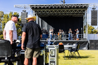 A band is performing on an outdoor stage with several musicians playing instruments, including guitars and drums. Two men in the foreground appear to be managing equipment, possibly part of the event staff. The stage is set up with a black backdrop and surrounded by speakers. The event seems to be taking place in a park or open field, with trees visible in the background.