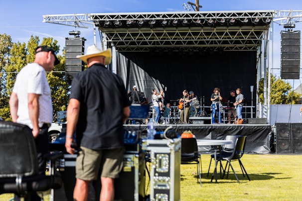 A band is performing on an outdoor stage with several musicians playing instruments, including guitars and drums. Two men in the foreground appear to be managing equipment, possibly part of the event staff. The stage is set up with a black backdrop and surrounded by speakers. The event seems to be taking place in a park or open field, with trees visible in the background.