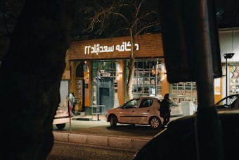 A street scene at night featuring a shop with illuminated signage in a language using Arabic script. In front of the shop, there are two parked cars and a person walking on the sidewalk. The shop's windows are lit and display various items. Trees and a dimly lit street add to the urban atmosphere.