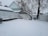 A snow-covered yard featuring a wooden fence and two leafless trees blanketed in snow. The area shows footprints in the snow, suggesting recent activity. In the background, there is a house with a triangular roof partially visible.