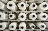 Multiple rolls of white fabric thread are stacked in an orderly fashion. The rolls are placed on a metal shelf, arranged vertically in rows. The perspective is from the top, showing the cylindrical shape of the rolls and the uniform pattern they create.