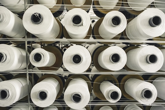 Multiple rolls of white fabric thread are stacked in an orderly fashion. The rolls are placed on a metal shelf, arranged vertically in rows. The perspective is from the top, showing the cylindrical shape of the rolls and the uniform pattern they create.