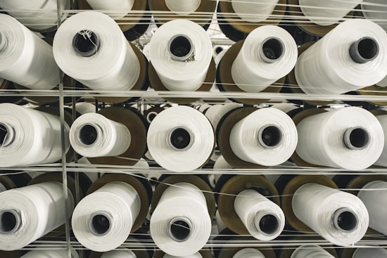 Multiple rolls of white fabric thread are stacked in an orderly fashion. The rolls are placed on a metal shelf, arranged vertically in rows. The perspective is from the top, showing the cylindrical shape of the rolls and the uniform pattern they create.