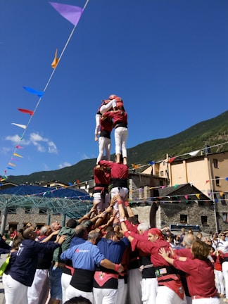 A group of people are building a human tower in an outdoor setting, surrounded by a festive atmosphere with colorful flags. The participants are wearing coordinated outfits, with those on the tower dressed in red shirts and white pants. The activity is taking place in a mountainous area with clear blue skies.