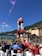 A group of people are building a human tower in an outdoor setting, surrounded by a festive atmosphere with colorful flags. The participants are wearing coordinated outfits, with those on the tower dressed in red shirts and white pants. The activity is taking place in a mountainous area with clear blue skies.