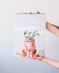 A person is holding up a spiral-bound calendar featuring the image of another person hiding their face behind a potted plant. The calendar cover has text in German, including the words 'immerwährender Pflanzen Kalender' and the brand name 'feey'. The background is plain and light-colored.