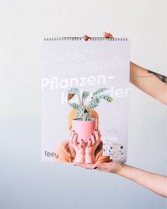 A person is holding up a spiral-bound calendar featuring the image of another person hiding their face behind a potted plant. The calendar cover has text in German, including the words 'immerw&auml;hrender Pflanzen Kalender' and the brand name 'feey'. The background is plain and light-colored.