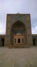 An ornate and detailed Islamic architectural structure with intricate geometric patterns and calligraphy. The colors are neutral, with a blend of light browns and blues. The entrance is arched with a pointed dome, typical of traditional mosque design.
