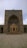 An ornate and detailed Islamic architectural structure with intricate geometric patterns and calligraphy. The colors are neutral, with a blend of light browns and blues. The entrance is arched with a pointed dome, typical of traditional mosque design.