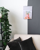 A minimalist interior setting featuring a white wall with a bold gray accent. A wall-mounted calendar with an image of a potted plant is displayed, and to the left, there is a green leafy plant cascading from a shelf. A beige sofa sits below, adorned with two dark gray pillows.