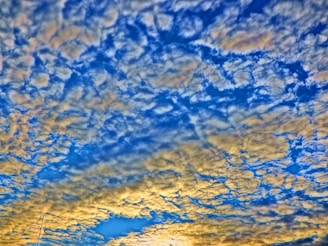 A vibrant scene featuring a dramatic cloud formation across the sky. The clouds have a textured appearance, with hues ranging from yellow to white, set against a bright blue background. A portion of an object resembling an antenna or structure can be seen in the lower left corner.