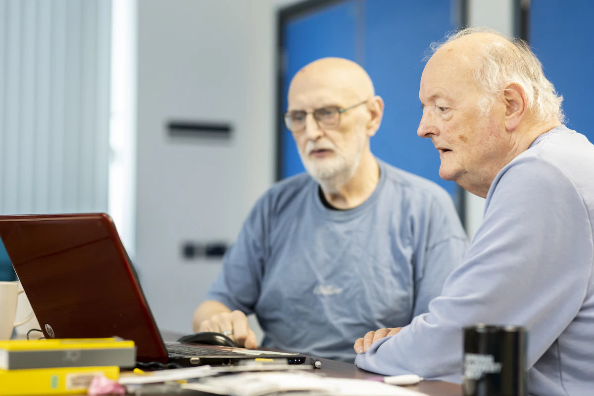 Deux hommes âgés assis côte à côte devant un ordinateur portable, l'un expliquant la manipulation à l'autre