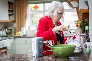 An older woman with white hair and glasses is in a kitchen, measuring flour with a spoon over a mixing bowl. She is wearing a bright red cardigan and a patterned top. The kitchen has a cozy and lived-in feel, with green cabinets, a fruit bowl on the counter, and a bright window letting in natural light. A mixer is on the counter alongside various containers of baking ingredients.