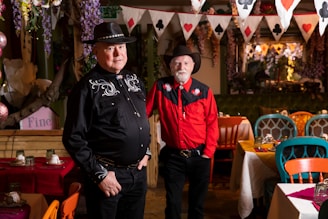 Two men wearing cowboy hats and western-style shirts stand in a colorful, decorated room with tables set for dining. The room features vibrant chairs and a festive string of playing card-themed flags hanging from the ceiling.