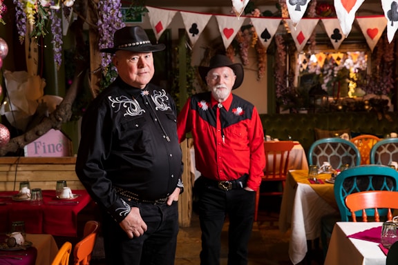 Two men wearing cowboy hats and western-style shirts stand in a colorful, decorated room with tables set for dining. The room features vibrant chairs and a festive string of playing card-themed flags hanging from the ceiling.