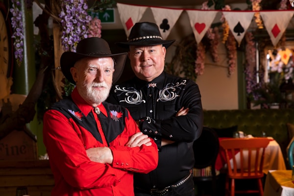 Two men dressed in Western-style clothing and cowboy hats are posing indoors. One is wearing a red shirt with black embroidered details and has his arms crossed, while the other is wearing a black shirt with white embroidery and has his hands folded. They are standing in front of a decorative background featuring hanging flowers and a string of bunting with card suits.