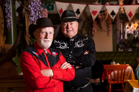 Two men dressed in Western-style clothing and cowboy hats are posing indoors. One is wearing a red shirt with black embroidered details and has his arms crossed, while the other is wearing a black shirt with white embroidery and has his hands folded. They are standing in front of a decorative background featuring hanging flowers and a string of bunting with card suits.