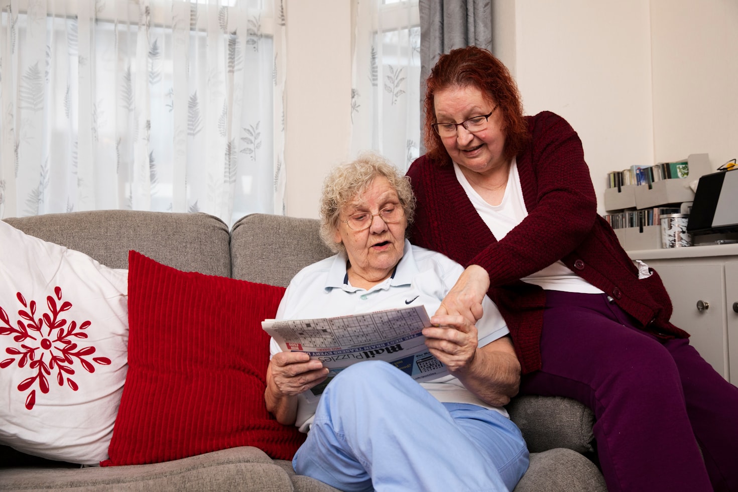 Two elderly women sitting on a couch reading a newspaper.