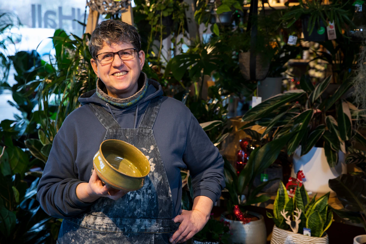 Potter in a clay-streaked apron smiles holding a yellow glazed bowl among plants.