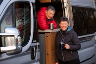 Couple brewing fresh coffee beside a serene loch with their camper van parked nearby at dawn.