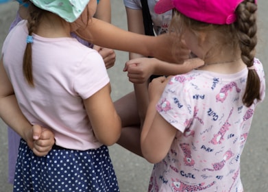 A group of children are standing close together, engaging in an activity. They are wearing colorful clothes and hats, and their arms are intertwined, indicating playfulness or a game.