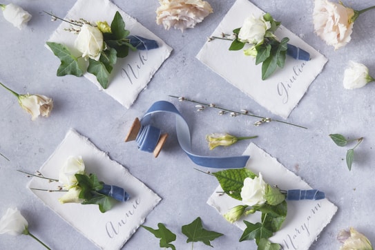 Elegant floral place cards are arranged on a textured gray surface. Each card is adorned with a small bouquet of white roses and green foliage, tied with a blue ribbon. The names Nik, Giulia, Annie, and Maryam are elegantly written on the cards. A wooden spool with blue ribbon lies nearby, surrounded by scattered flowers and leaves.