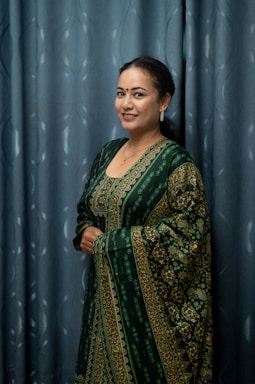 A woman wearing a traditional green and gold patterned dress stands against a blue curtain backdrop. She has a smile on her face, a bindi on her forehead, and is accessorized with earrings and a necklace. Her hands are held together in front of her, and her hair is styled back.